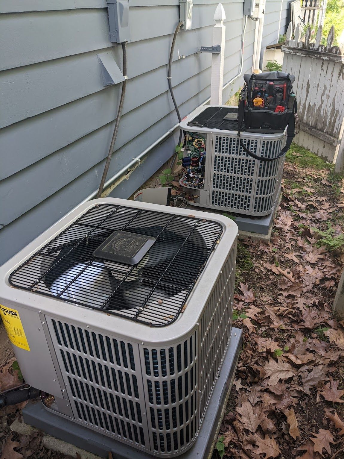 Two grey outdoor HVAC condenser units sitting on blocks near a house siding, with a tool bag on the back unit.