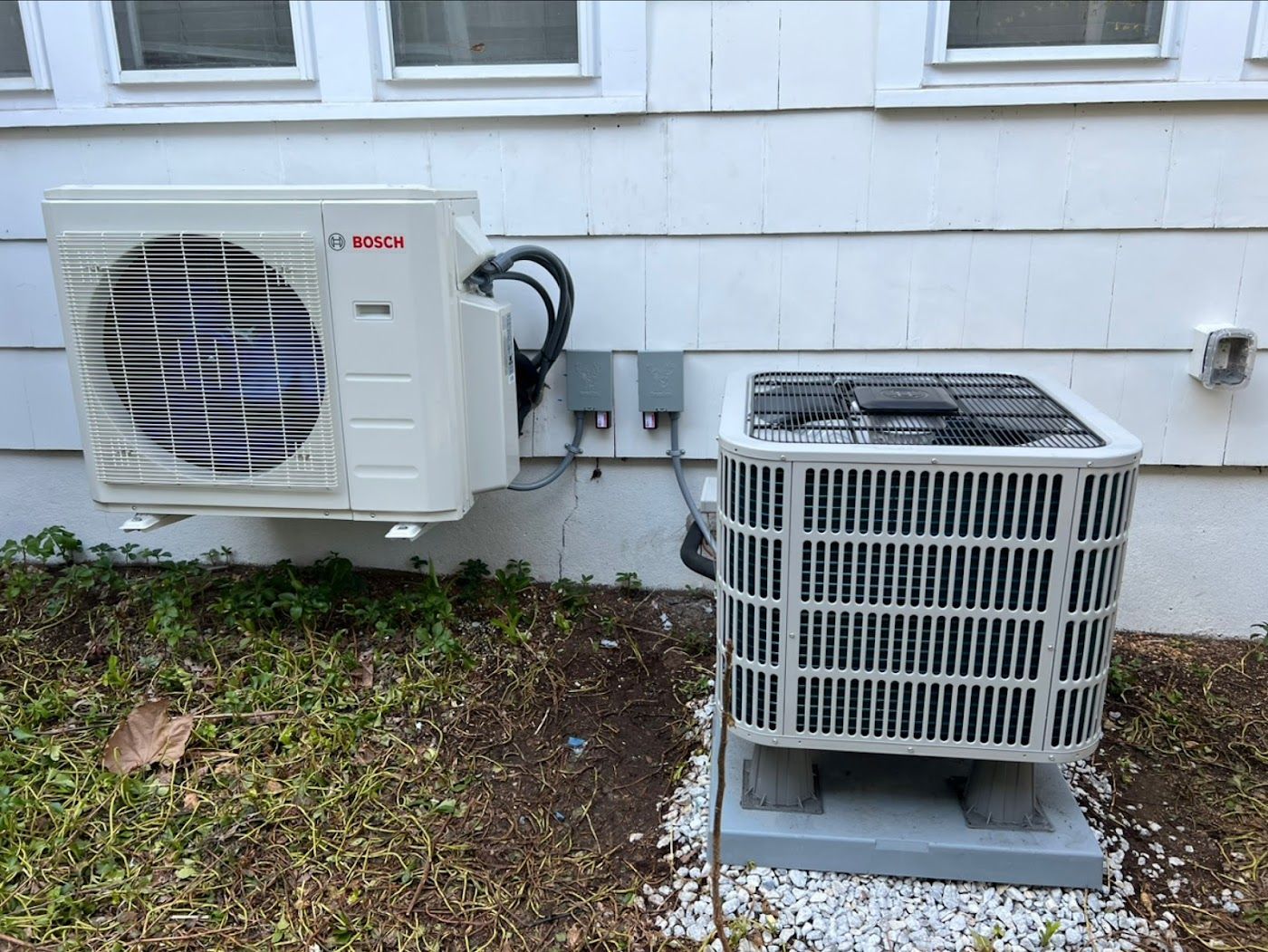 Two outdoor HVAC units, one wall-mounted and one ground-mounted, positioned against a white house siding.
