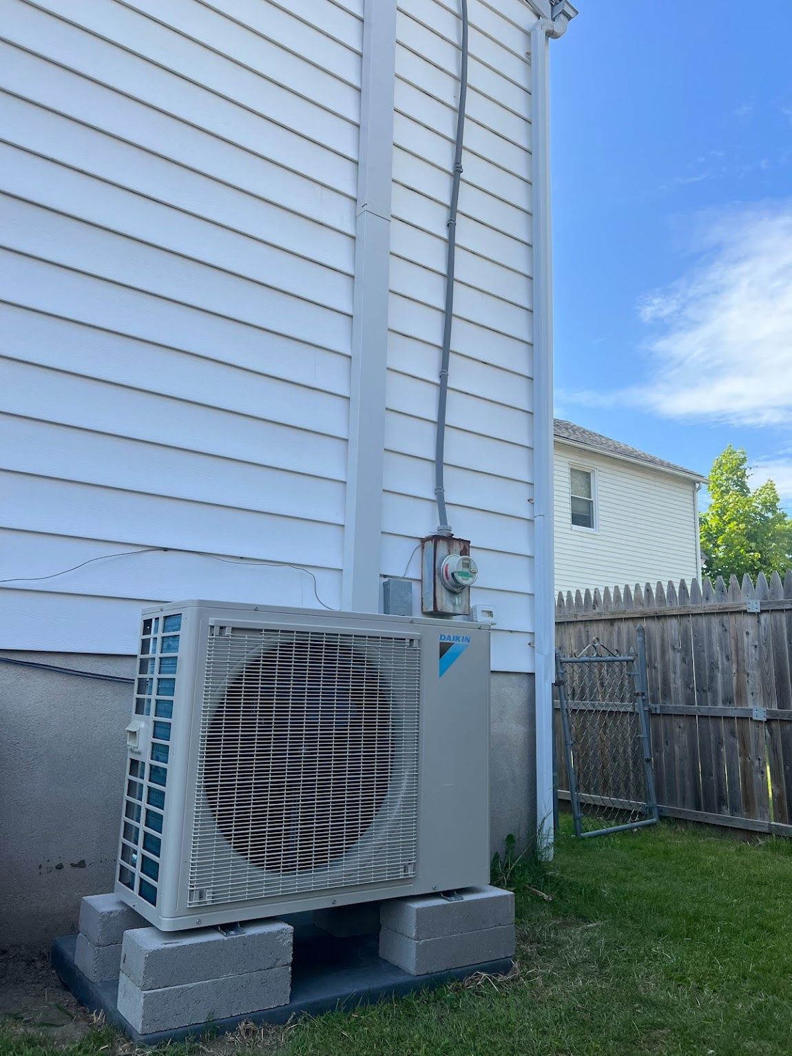 A Daikin outdoor HVAC unit sits on concrete blocks against the white-sided wall of a house with a wooden fence nearby.