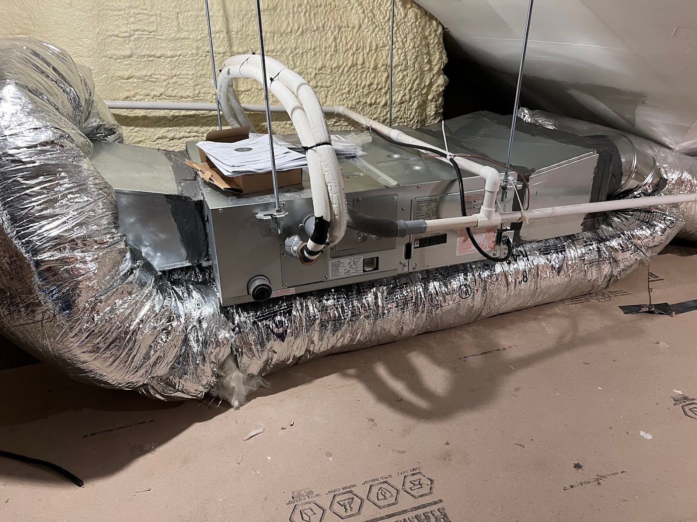 An HVAC air handler suspended in an attic with insulated ductwork and a white condensate drain line.