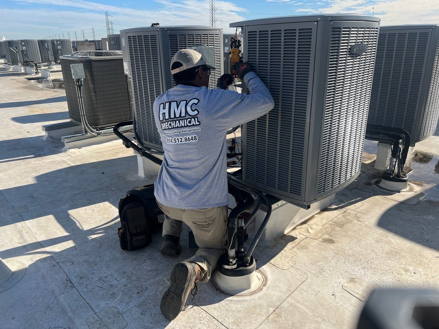 A technician in a gray shirt kneeling on a rooftop, working on the side of a large commercial HVAC unit.