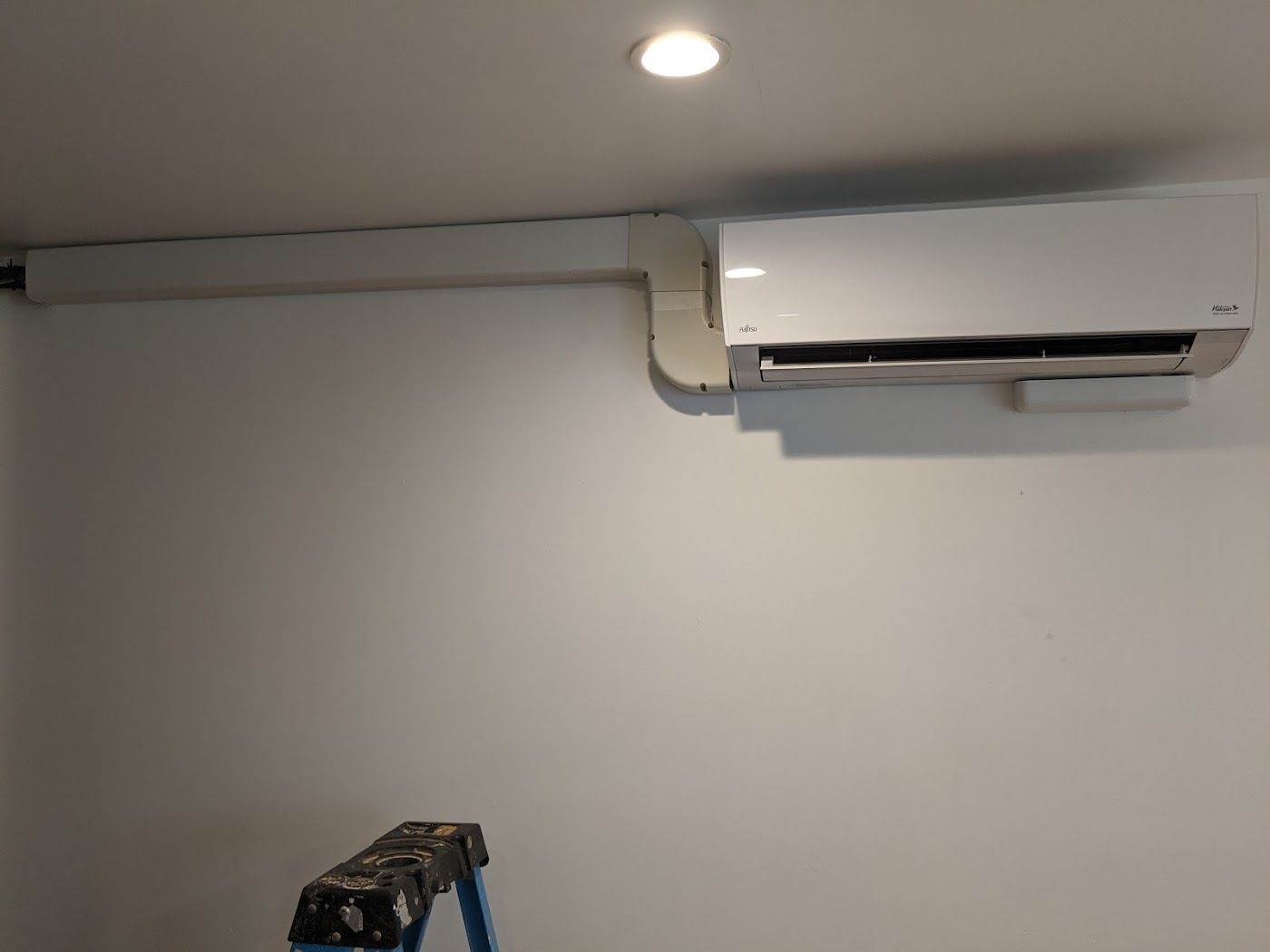 A white duct cover runs along the wall toward a wall-mounted indoor air conditioning unit inside a room.