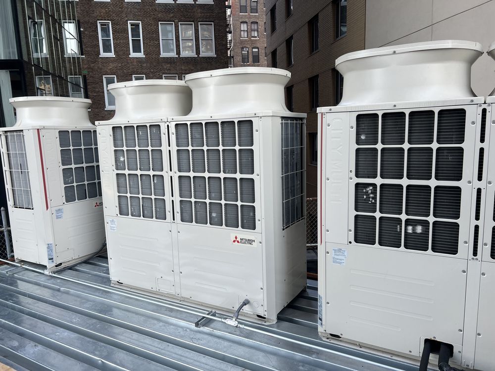 Several white HVAC units stand on a corrugated metal rooftop against a backdrop of city buildings.