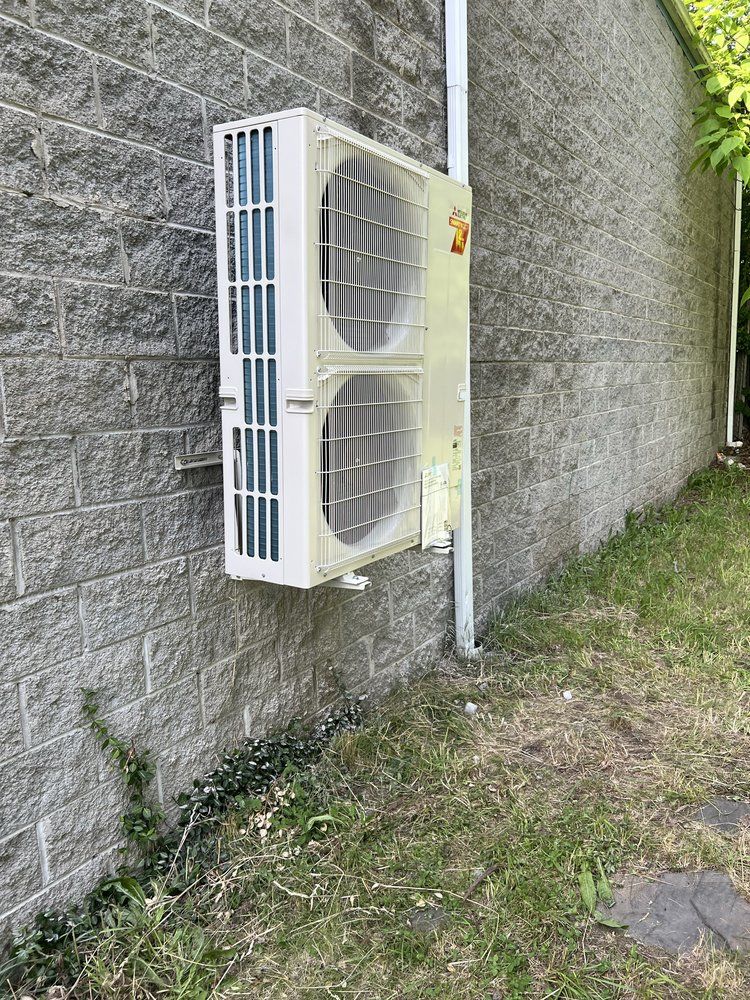 A white, dual-fan HVAC condenser unit mounted on a textured gray concrete block wall outdoors.