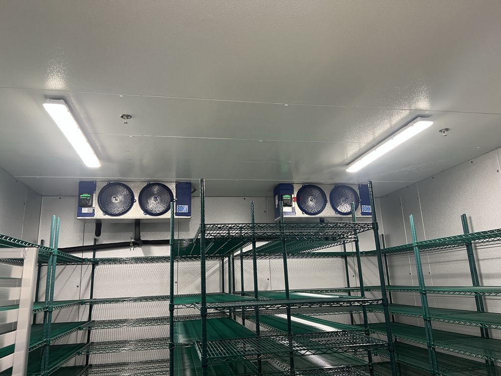 A commercial walk-in cooler or freezer with ceiling lights, two industrial fan units, and empty green metal shelving.