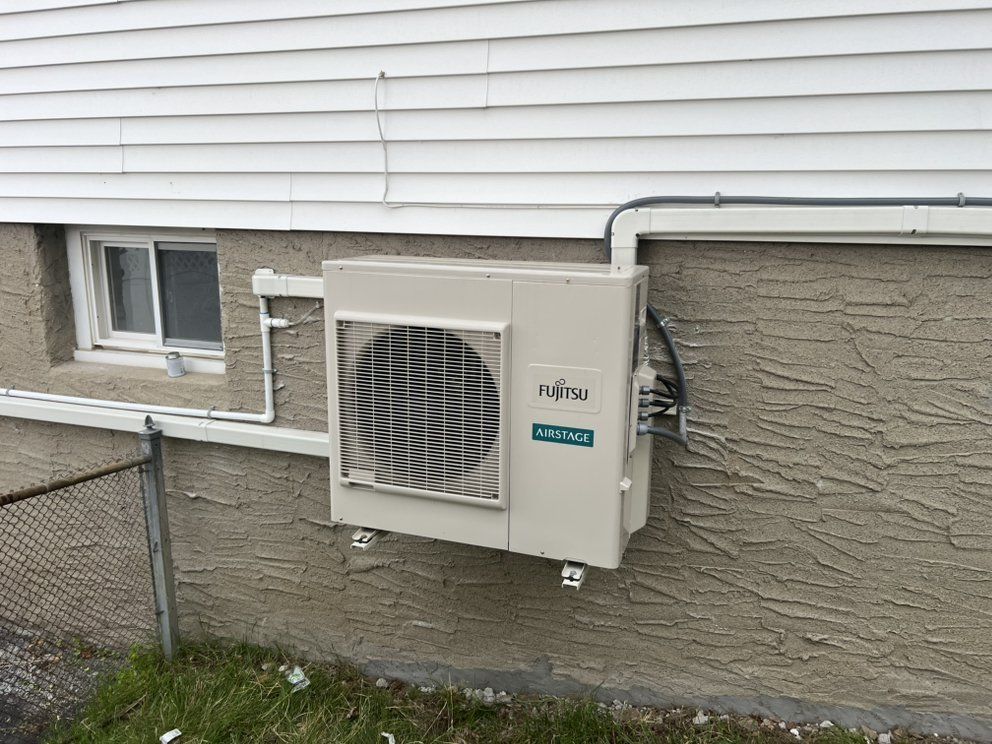 A wall-mounted Fujitsu outdoor HVAC unit attached to the side of a house with white siding and a stucco foundation.