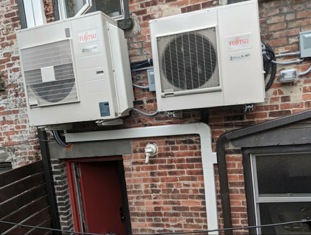 Two Fujitsu air conditioning units mounted on a brick exterior wall above a red doorway and a window.