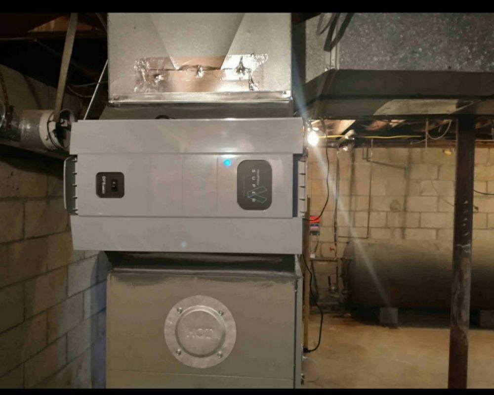 A grey HVAC air filtration unit mounted on a furnace in a basement with a cinder block wall and a large tank in the rear.