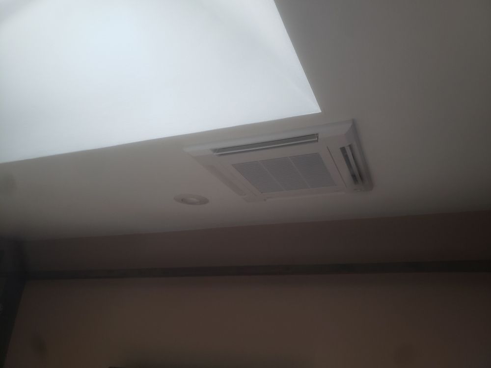 A square, white ceiling-mounted HVAC unit installed in a smooth, white ceiling near a skylight.