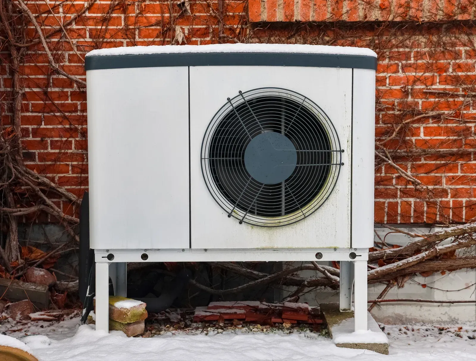 An outdoor heat pump unit sits on a snowy ground in front of a red brick wall covered with vines.