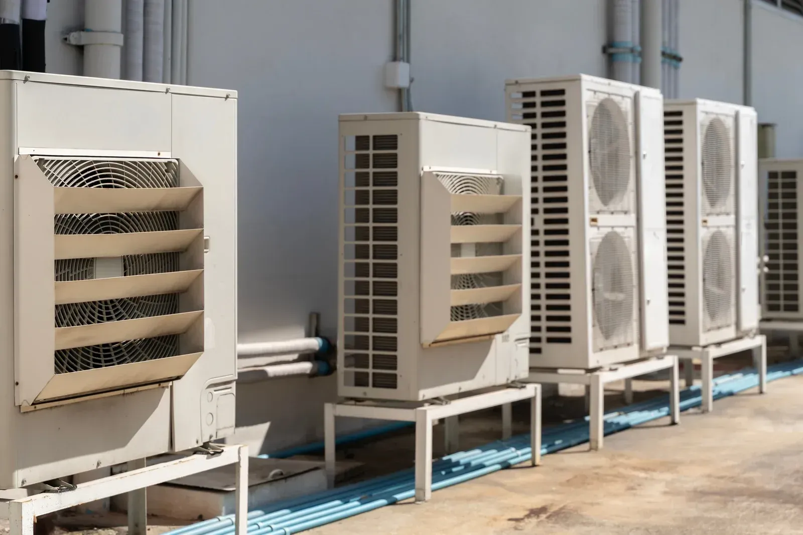 A row of industrial beige air conditioning units standing on a concrete outdoor surface against a white wall.