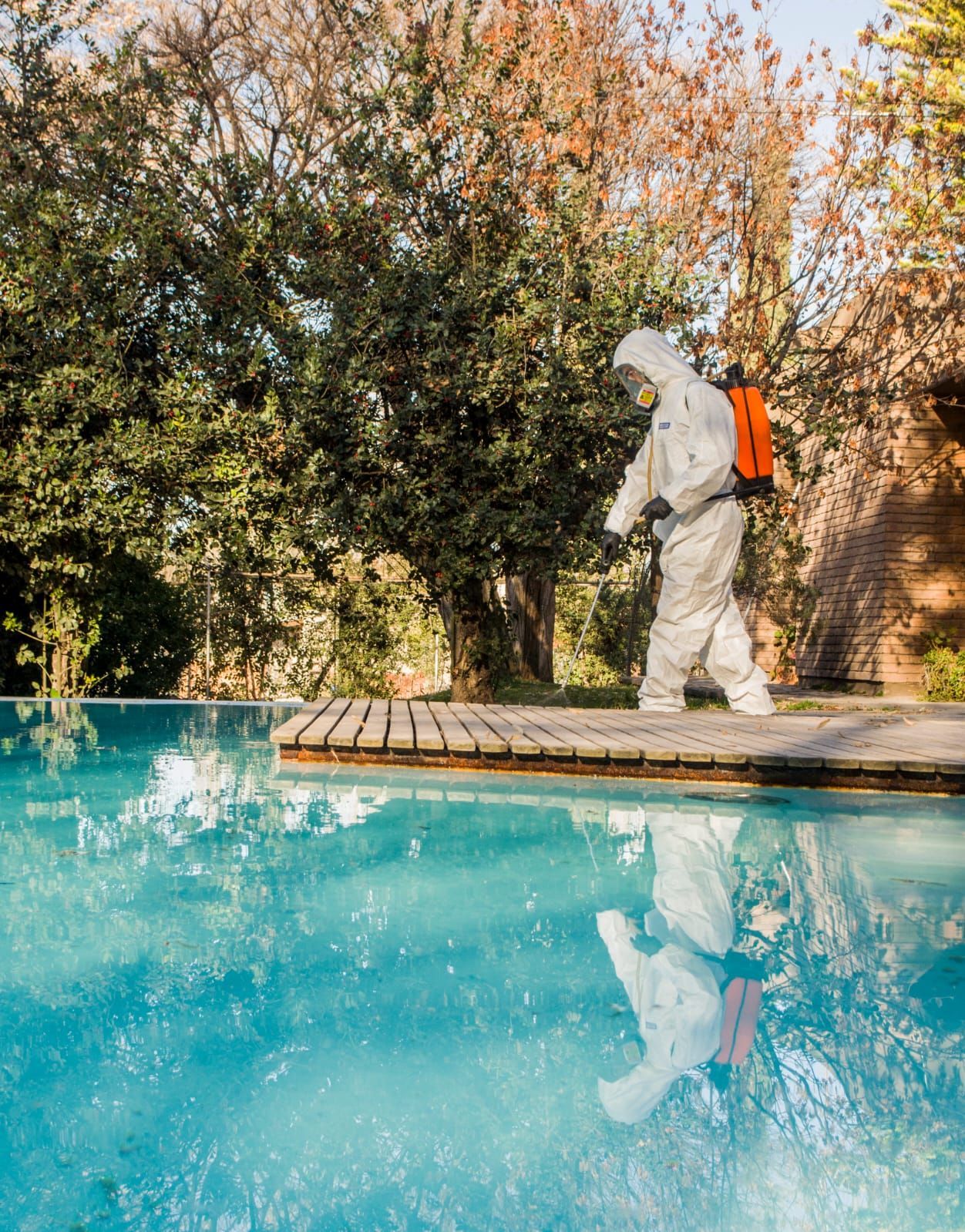 Una persona con traje de materiales peligrosos desinfecta la piscina con árboles de fondo.