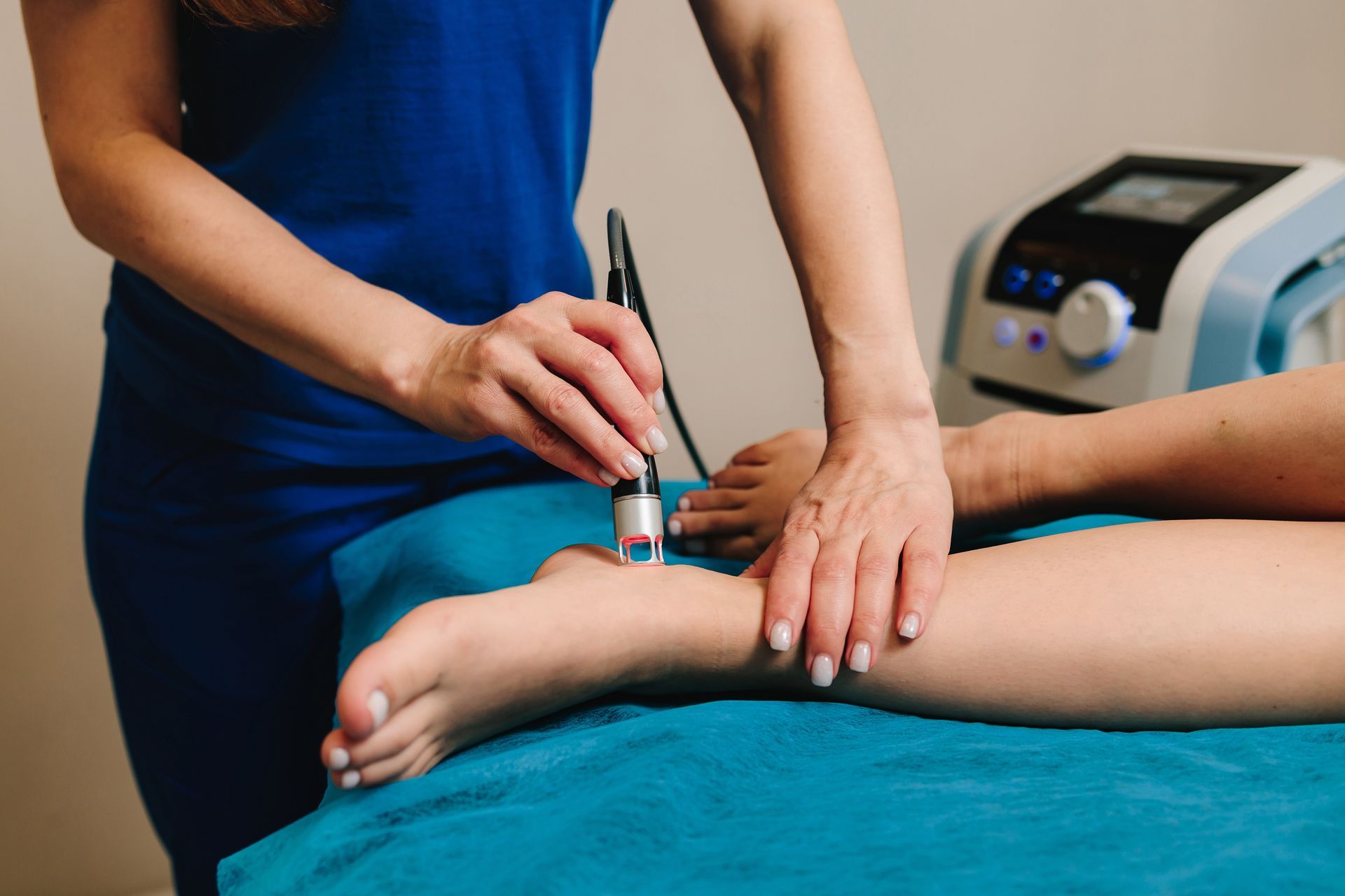 Therapist using a medical device on a patient's ankle, indoors. Blue and white equipment, turquoise table.