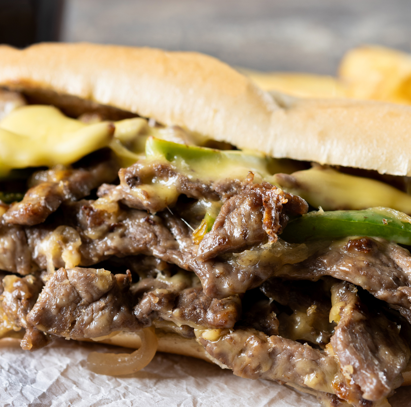 A close up of a steak and cheese sandwich with french fries in the background.