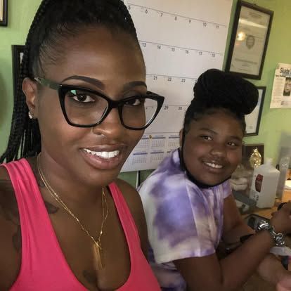 Two women smiling for the camera. and a pink top. The other has a purple tie-dye shirt and a bun. They are indoors.