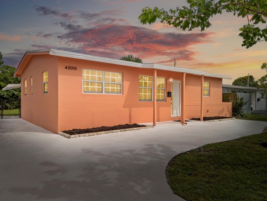 Front exterior of a single-story coral-colored home at 4200 NW 12th Avenue, Oakland Park, Florida, with a wide driveway, white trim, and sunset sky in the background.