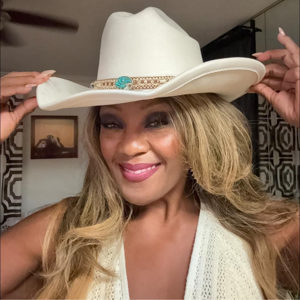 Woman with long blonde hair wearing a white cowboy hat, smiling, holding the hat with her hands.