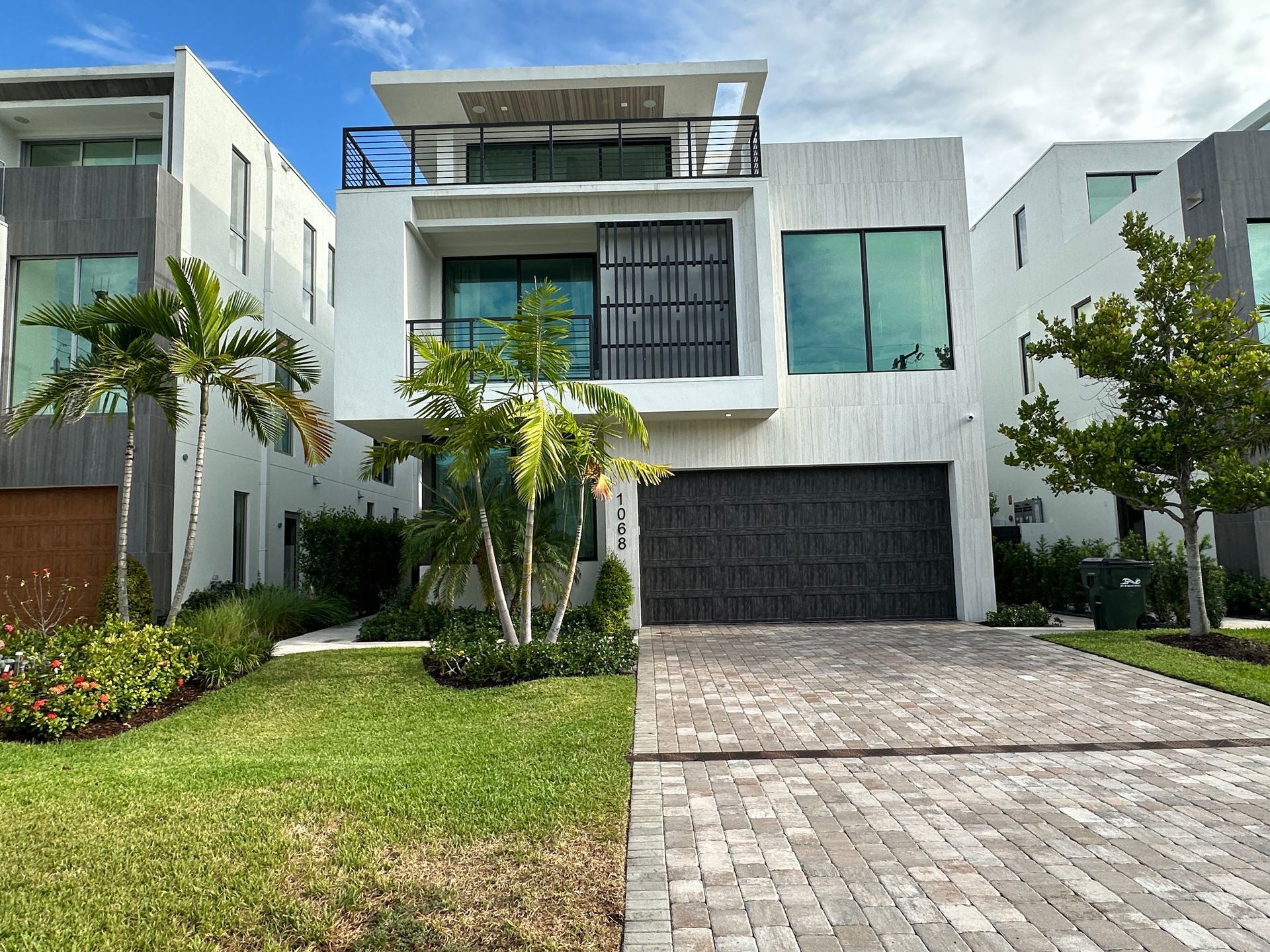 Modern white stucco house with black garage door and brick driveway; sunny day.