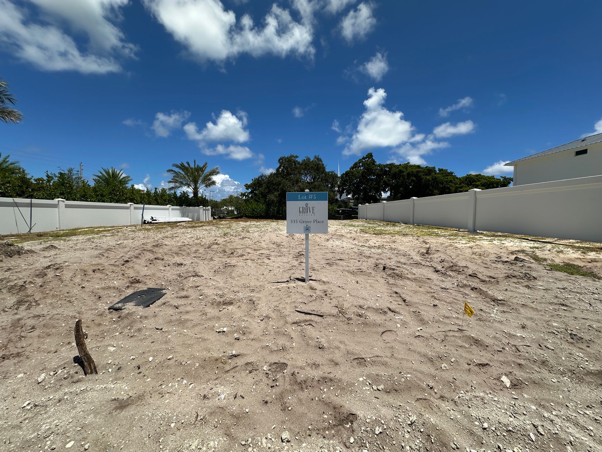 Empty lot with a sign against a blue sky with scattered clouds, enclosed by white walls and trees.