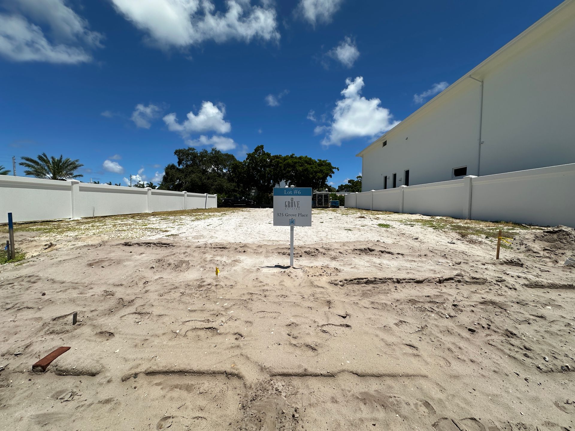 A vacant lot with a sign on a sunny day. Sand, white walls, blue sky, and sparse vegetation are present.