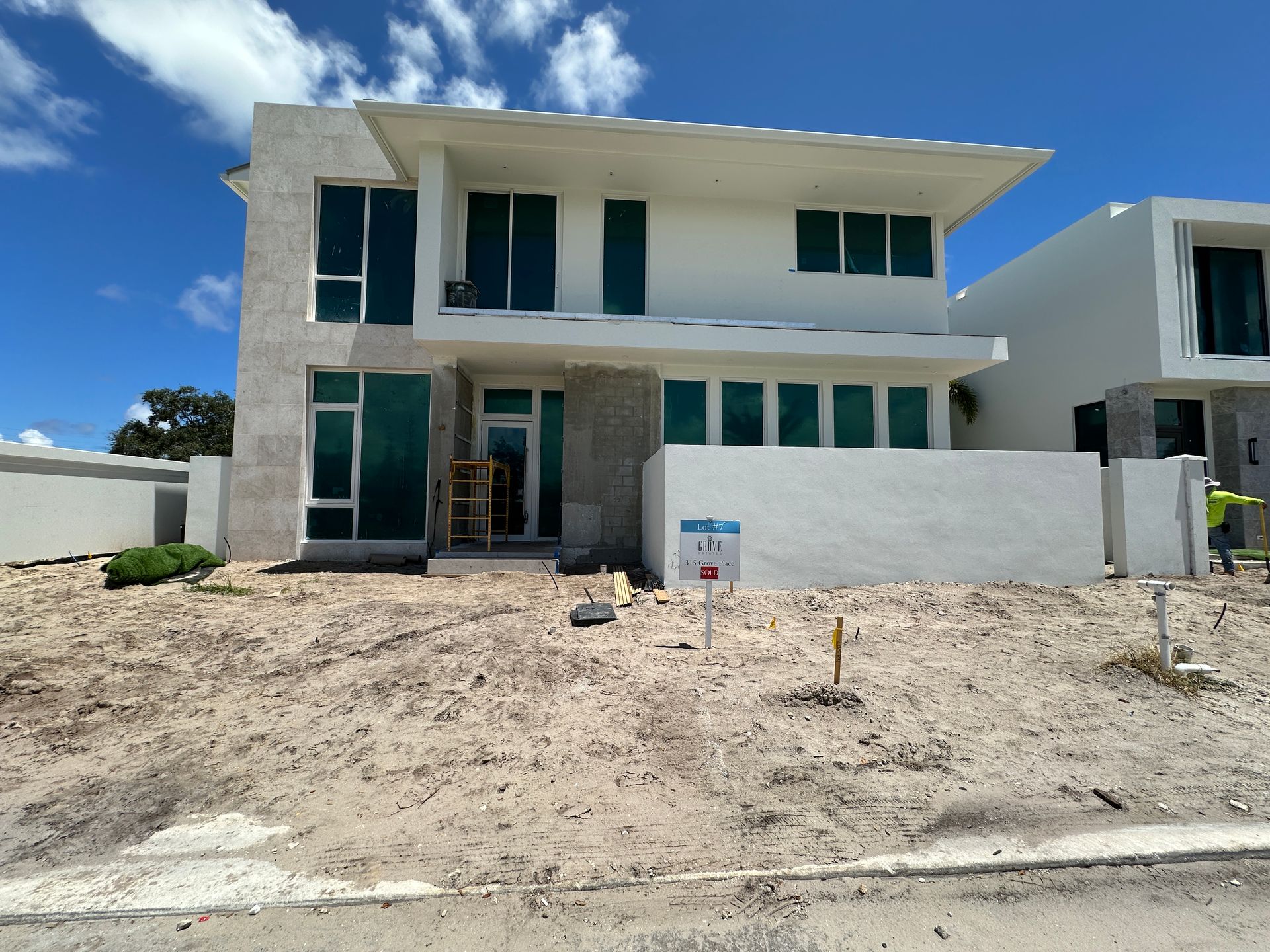Modern two-story house with large windows, light-colored facade, and construction site setting. Blue sky overhead.