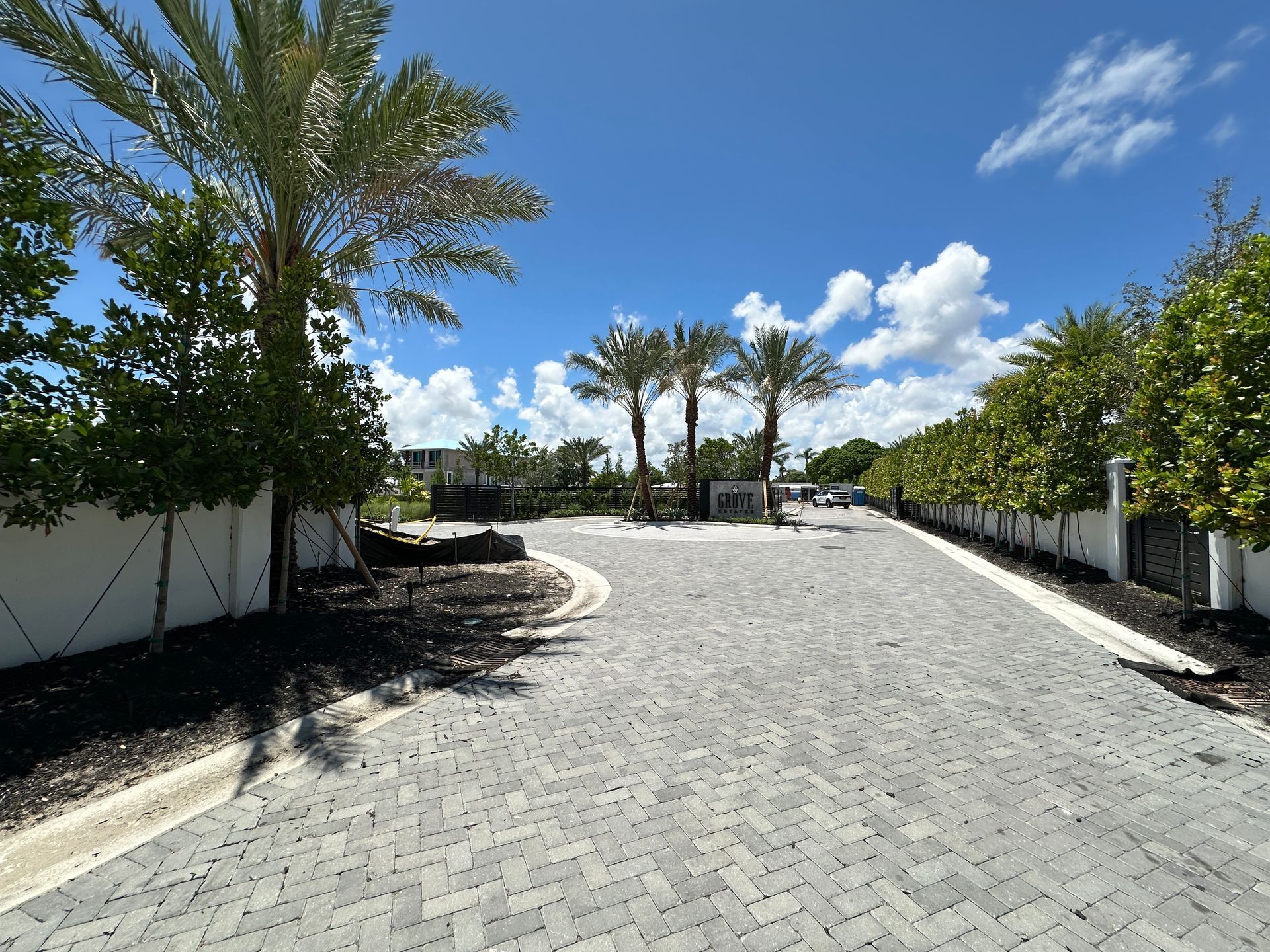 Brick road leading to a gated entrance, lined with trees and palm trees against a bright blue sky.