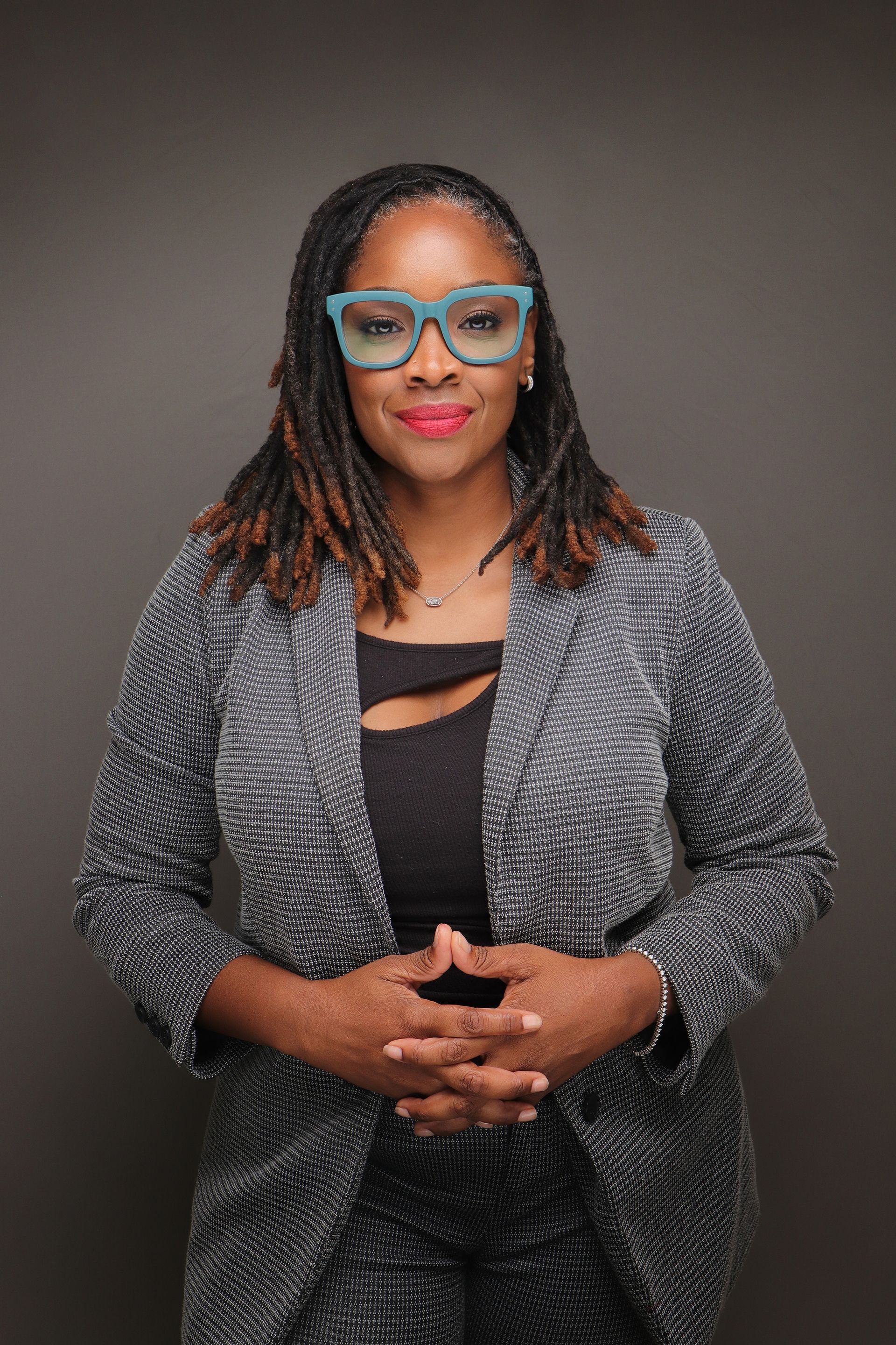 Woman in glasses, business suit, hands clasped, smiling, against a gray backdrop.