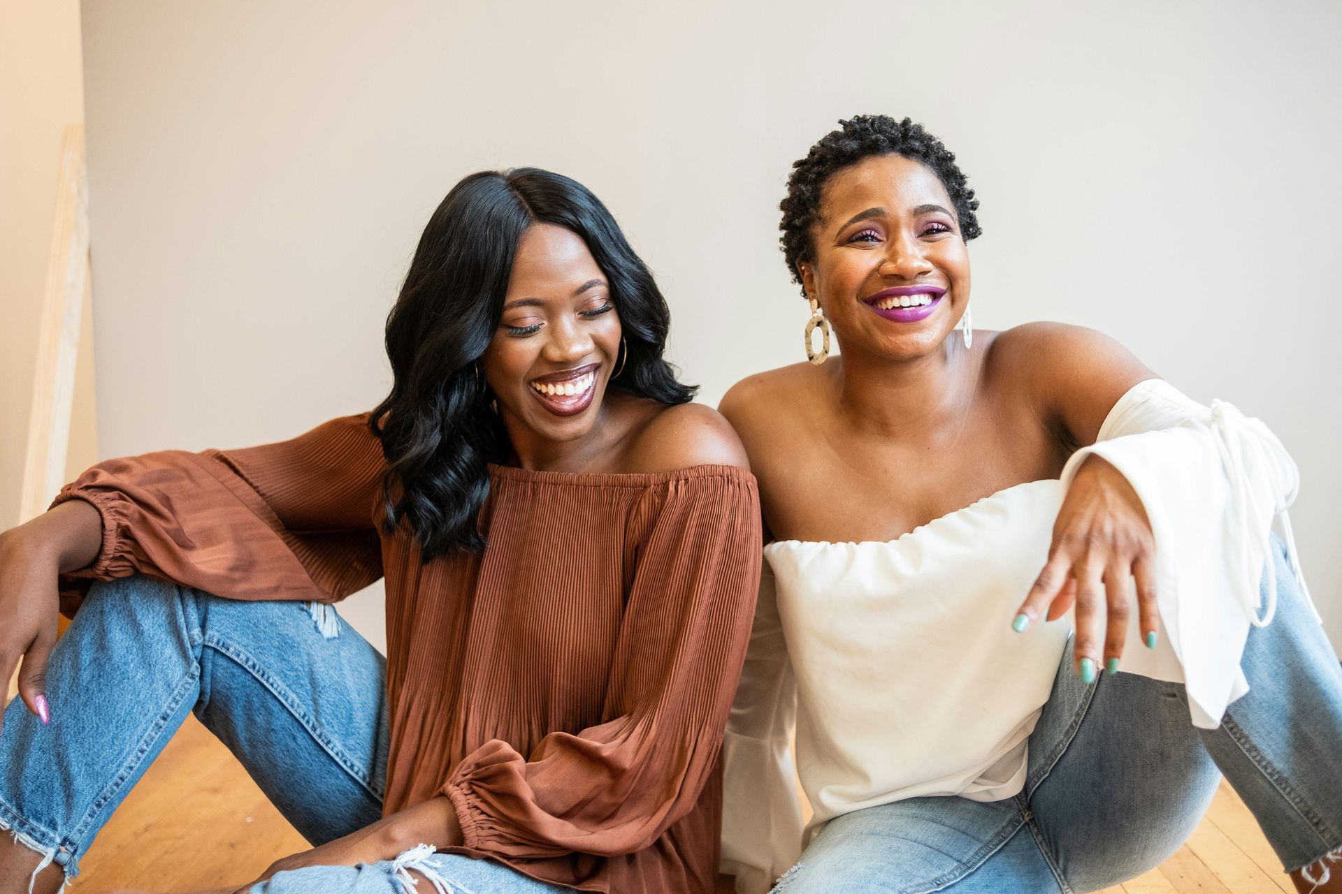Two smiling Black women sit together indoors. One wears a brown off-shoulder top, the other a white off-shoulder top, both in jeans.