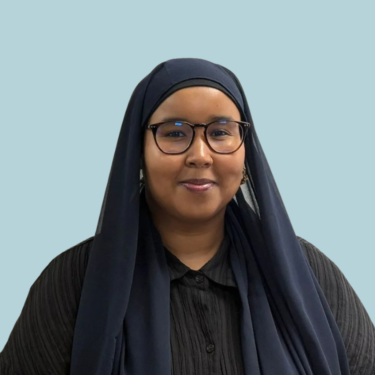 Woman with glasses and dark headscarf, smiling, against a light blue background.