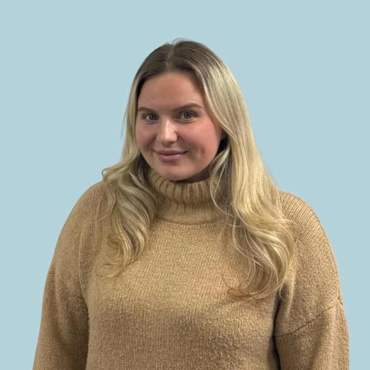 Woman with long blonde hair wearing a tan turtleneck sweater, smiling, against a light blue backdrop.