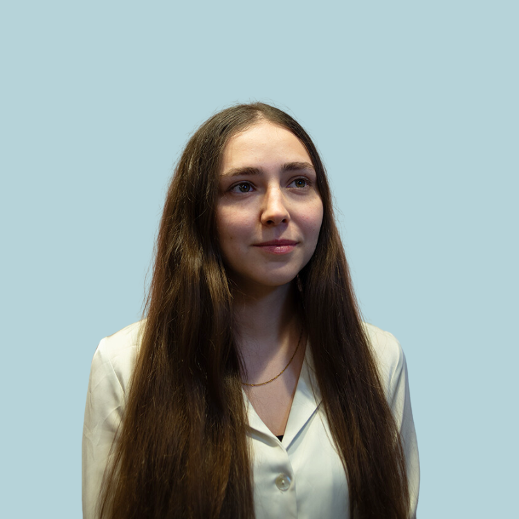 Woman with long brown hair, wearing a white button-up shirt, looking to the side. Light blue background.