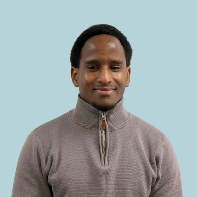 Man with dark hair and a tan sweater smiles, chest up, against a light blue background.