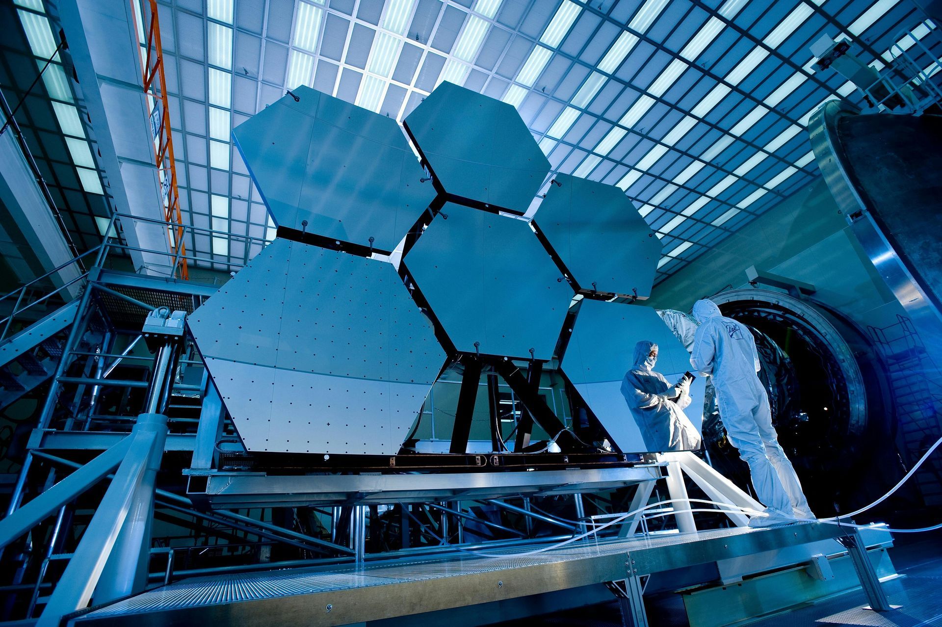 Hexagonal mirror segments in a laboratory; a technician in a white suit works on them.