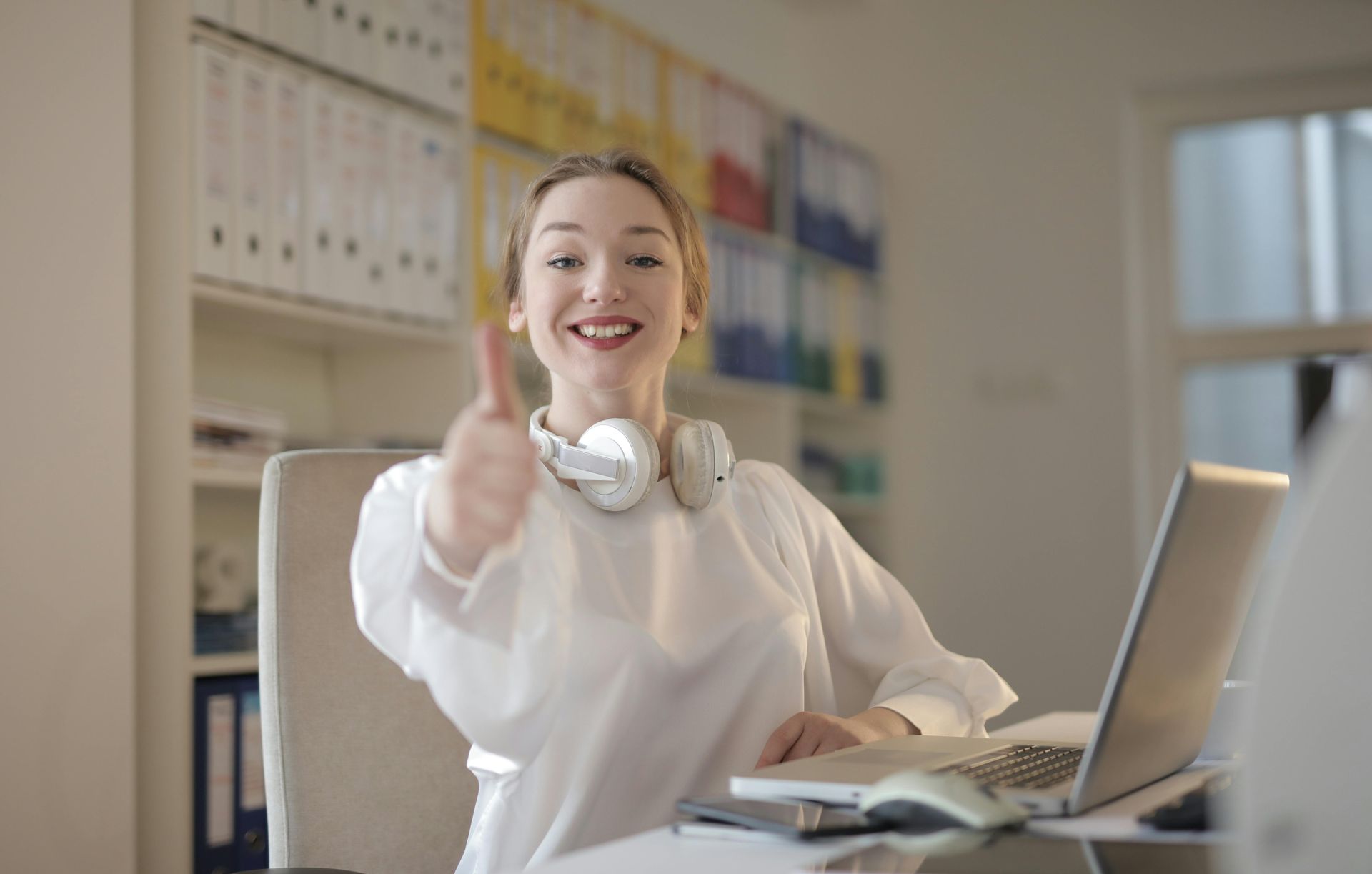 Woman at desk gives thumbs up, smiling at laptop, headphones around neck.