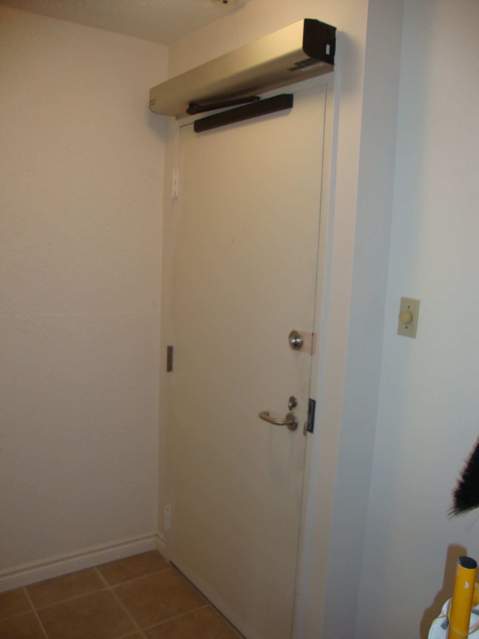 Porte blanche avec ouvre-porte automatique, poignée et serrure, dans un couloir aux murs blancs.