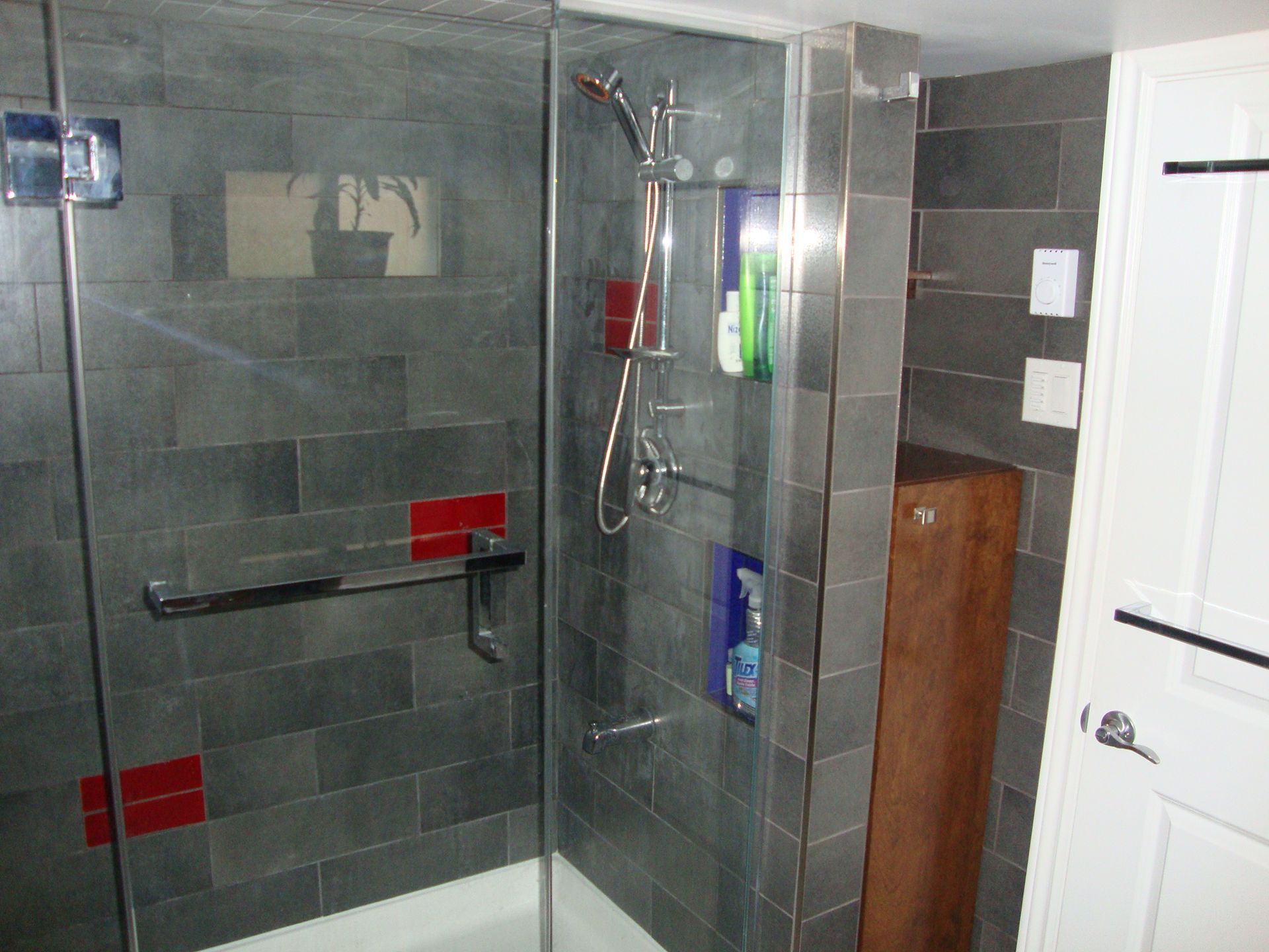Douche carrelée grise avec porte en verre, robinetterie argentée, touches de rouge et meuble en bois.