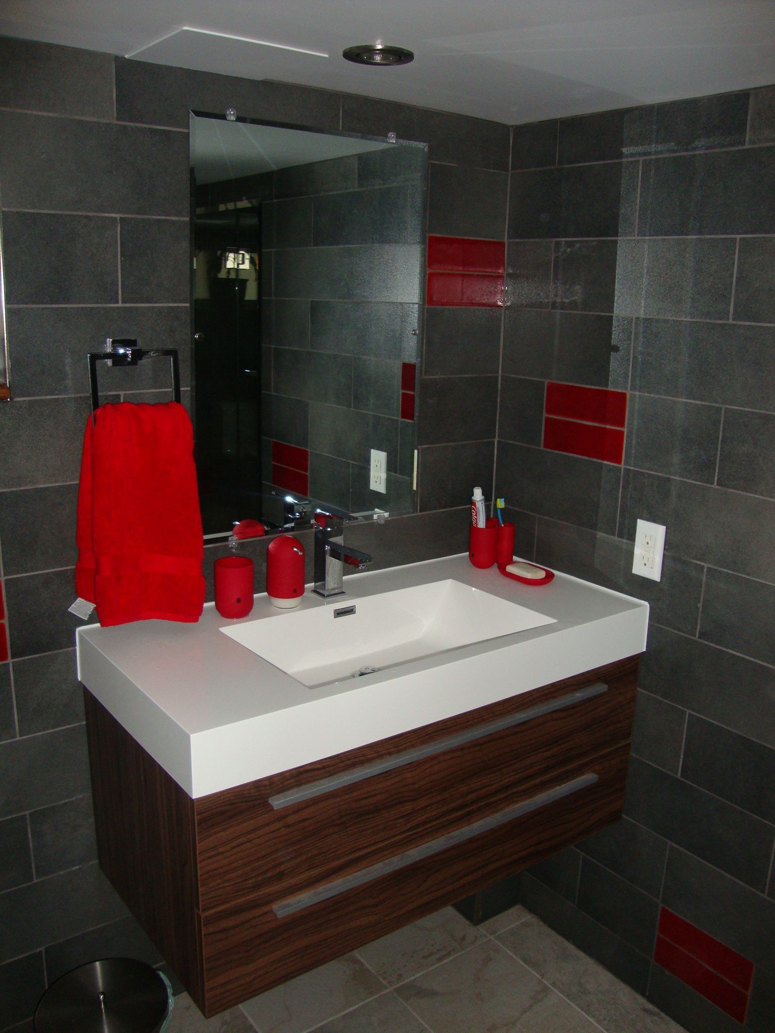 Salle de bains moderne avec murs carrelés gris, touches de rouge et meuble-lavabo en bois.