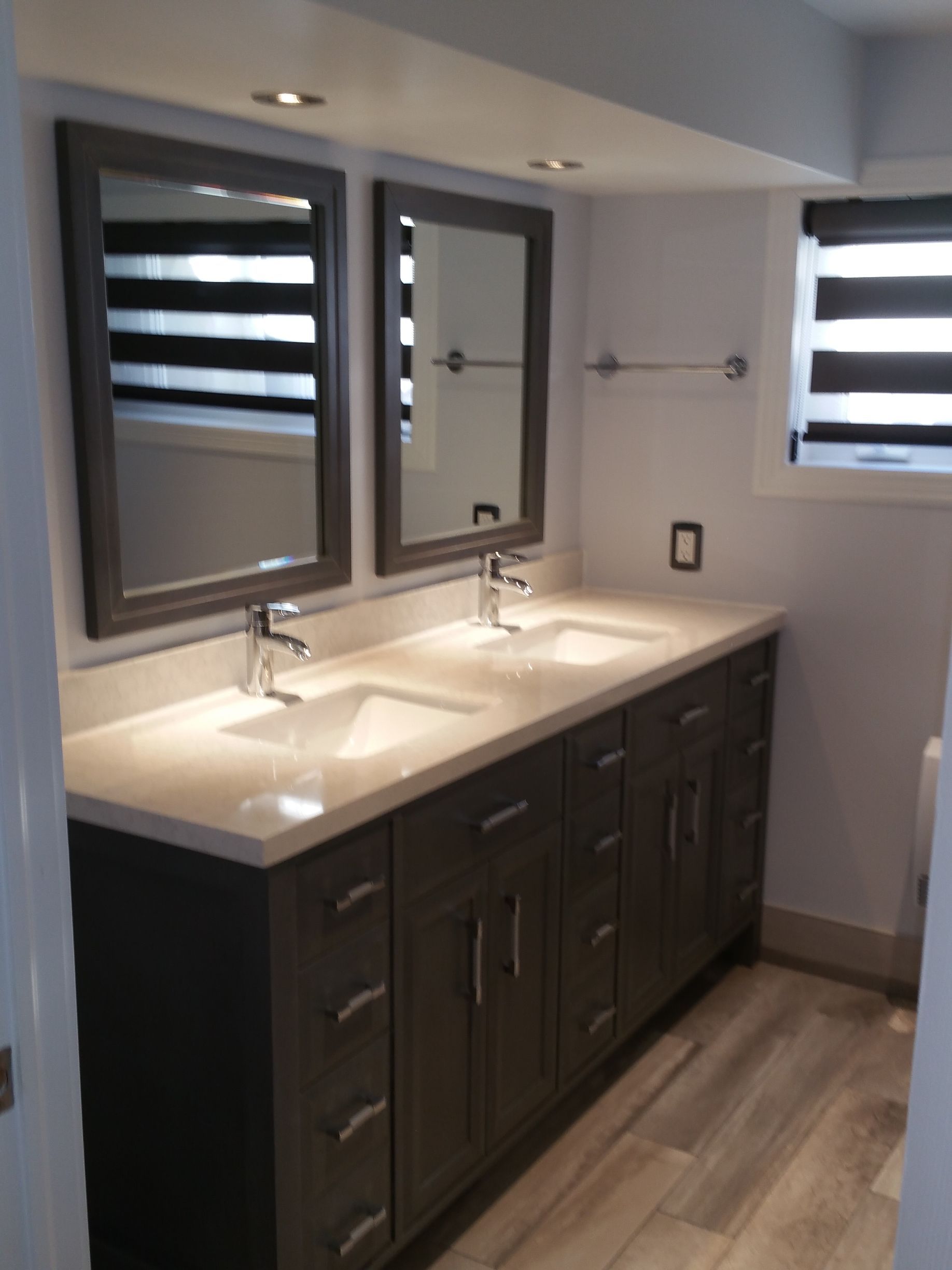 Salle de bains avec double vasque. Deux miroirs, meubles gris, plan de travail clair et sol imitation bois.