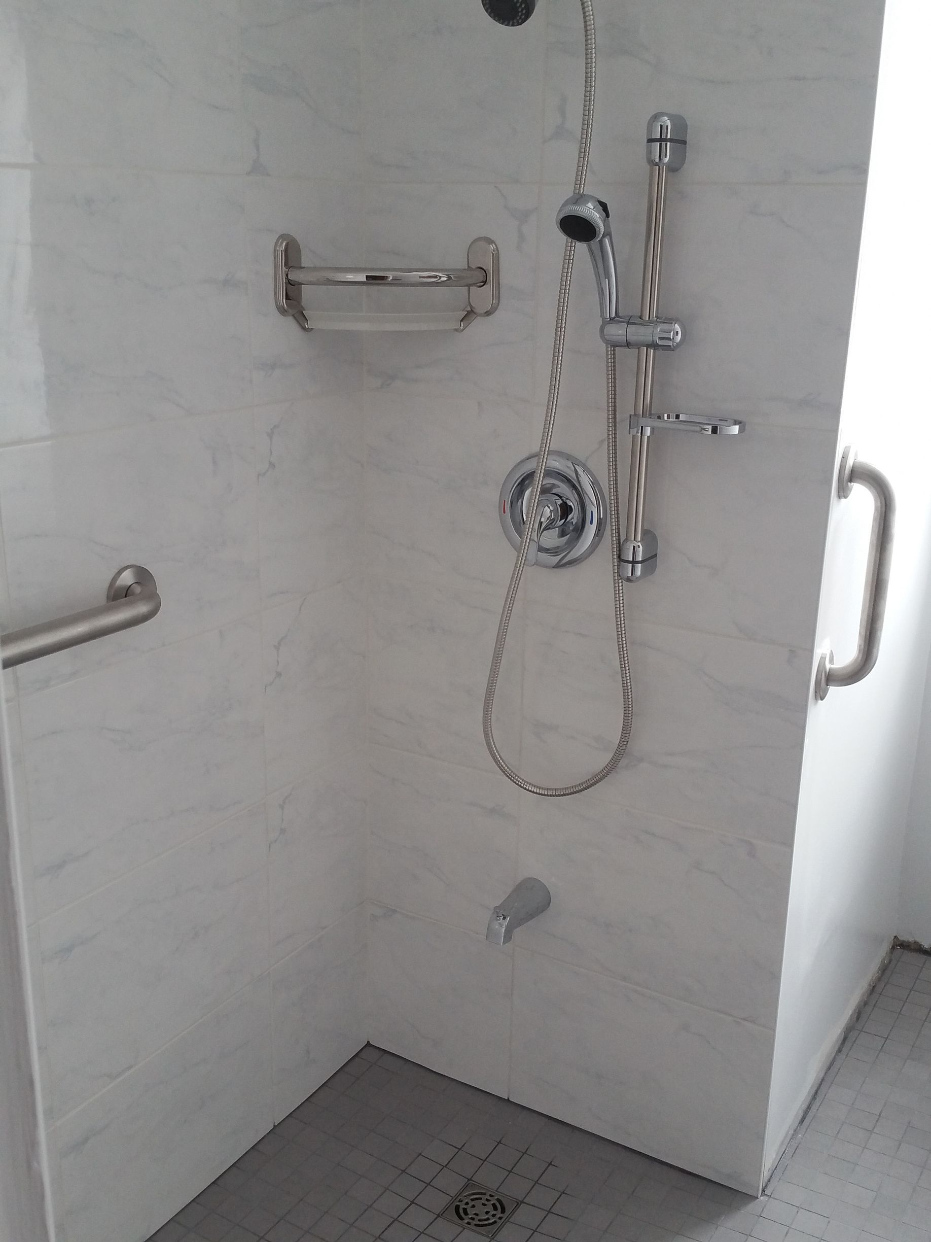 Douche accessible avec barres d'appui et douchette, carrelage en marbre blanc, receveur de douche gris et robinetterie chromée.