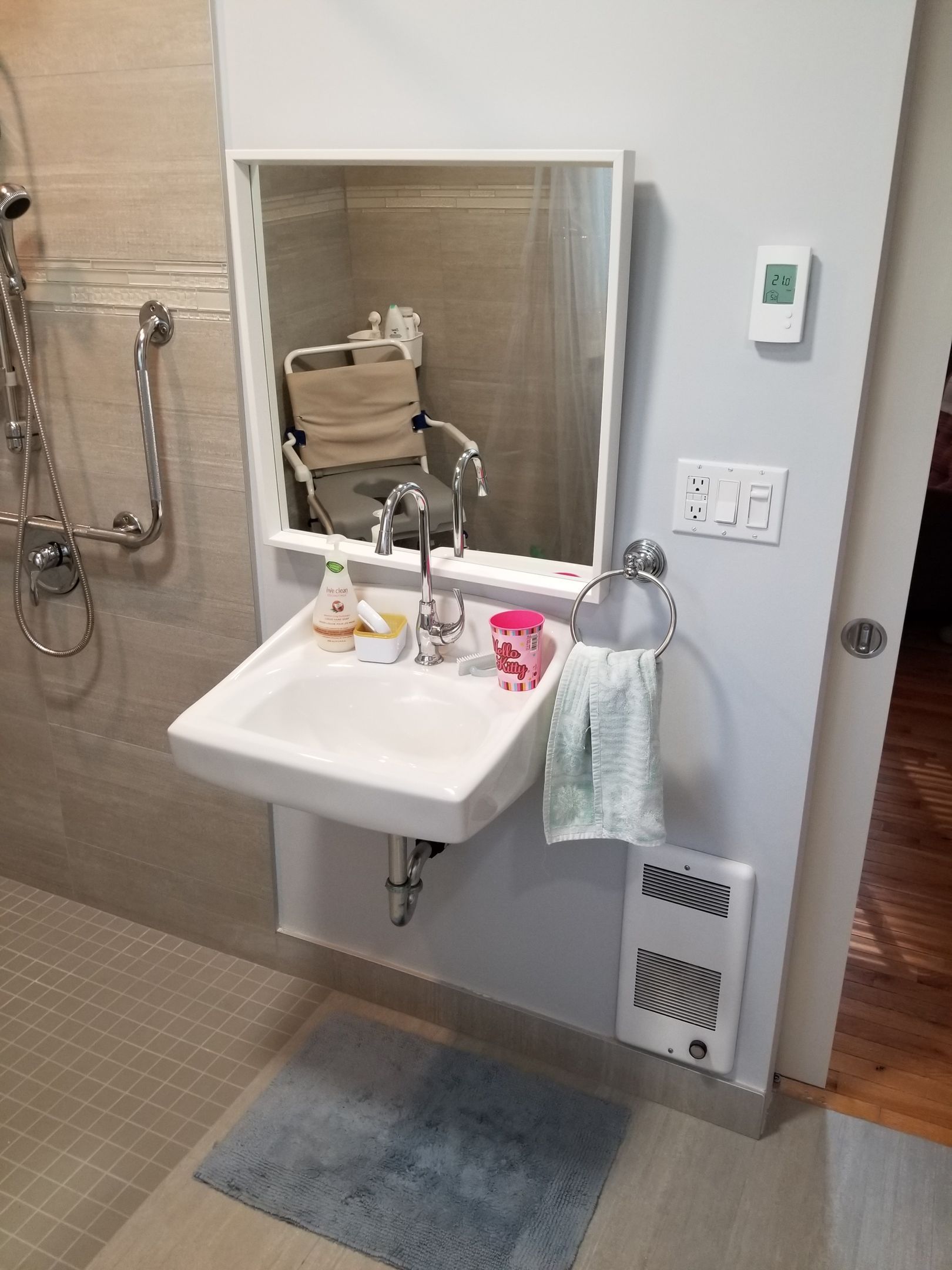 Salle de bains avec lavabo mural, miroir, douche, barre d'appui, porte-serviettes, tapis et chauffage. Tons beige clair et bleu.