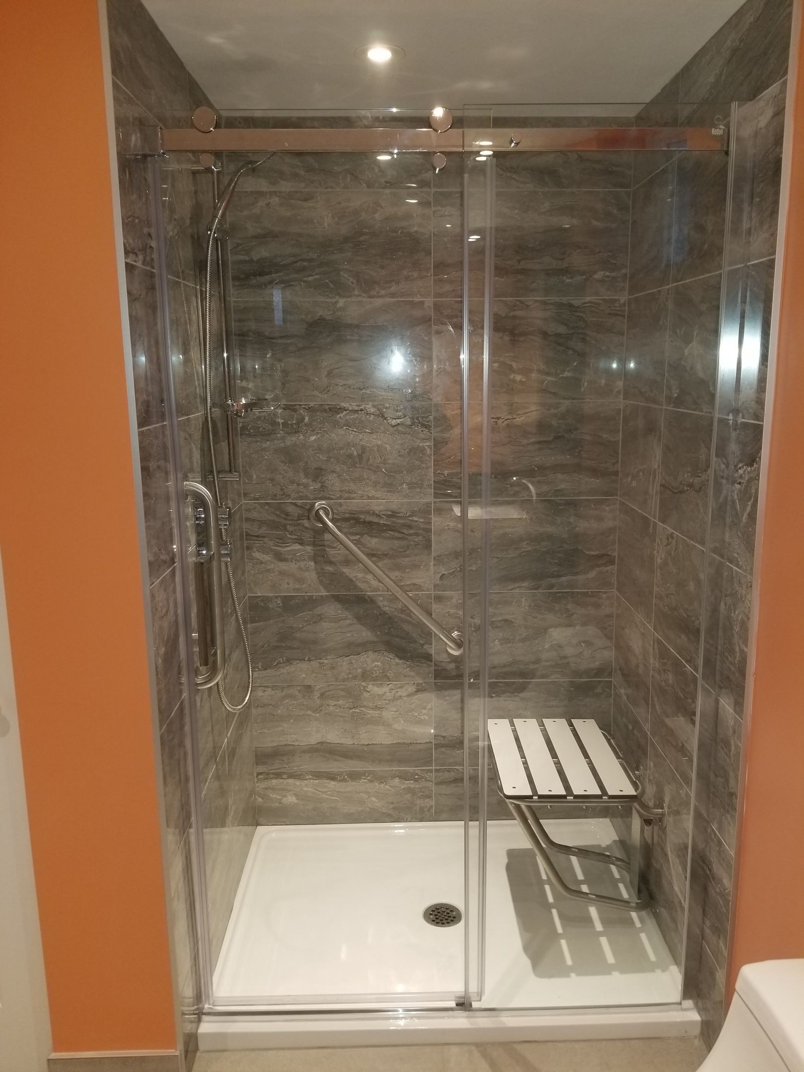 Une douche à l'italienne avec carrelage gris, portes coulissantes en verre, siège blanc et barre d'appui.