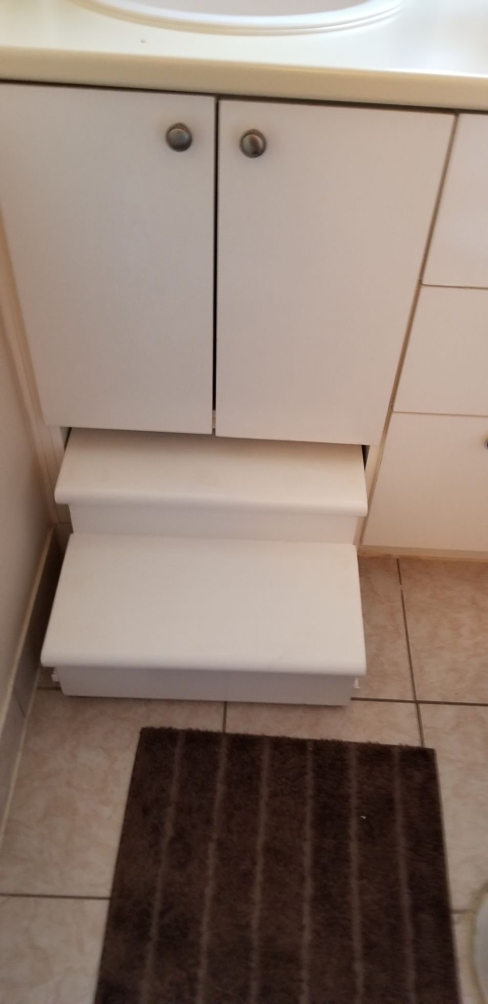 Meuble blanc avec marchepied escamotable. Marchepied rétracté. Tapis de bain marron sur carrelage.