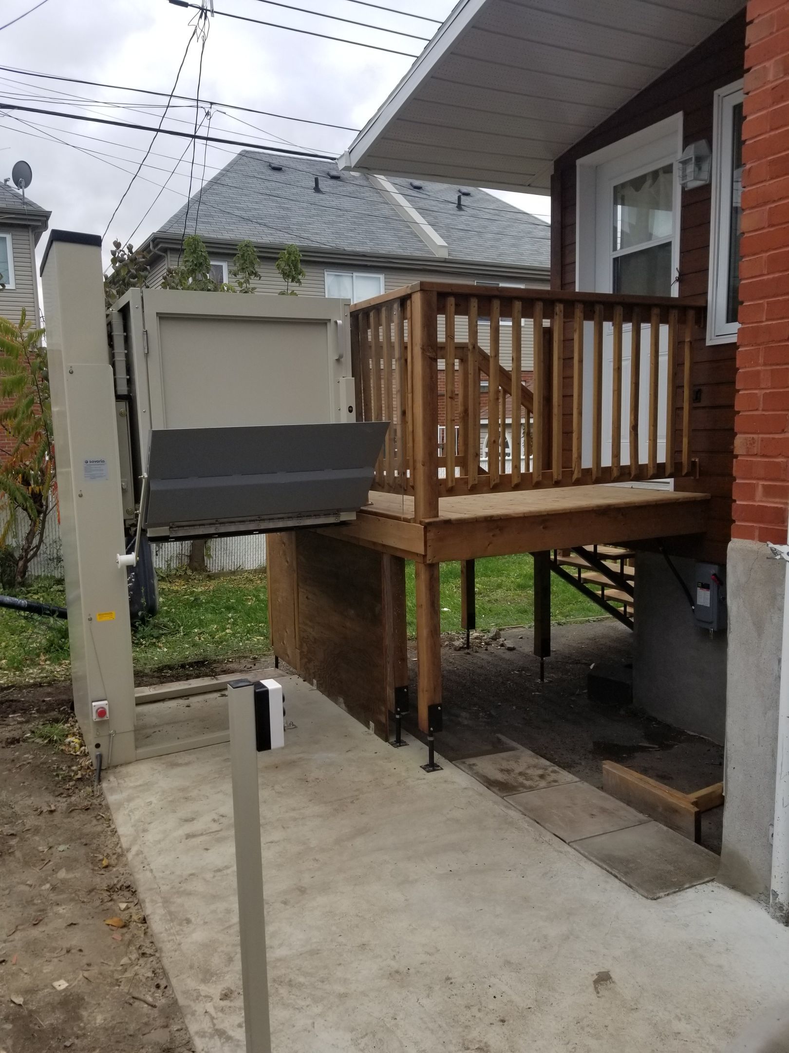 Un ascenseur vertical extérieur, à côté d'une terrasse en bois attenante à un bâtiment en briques.