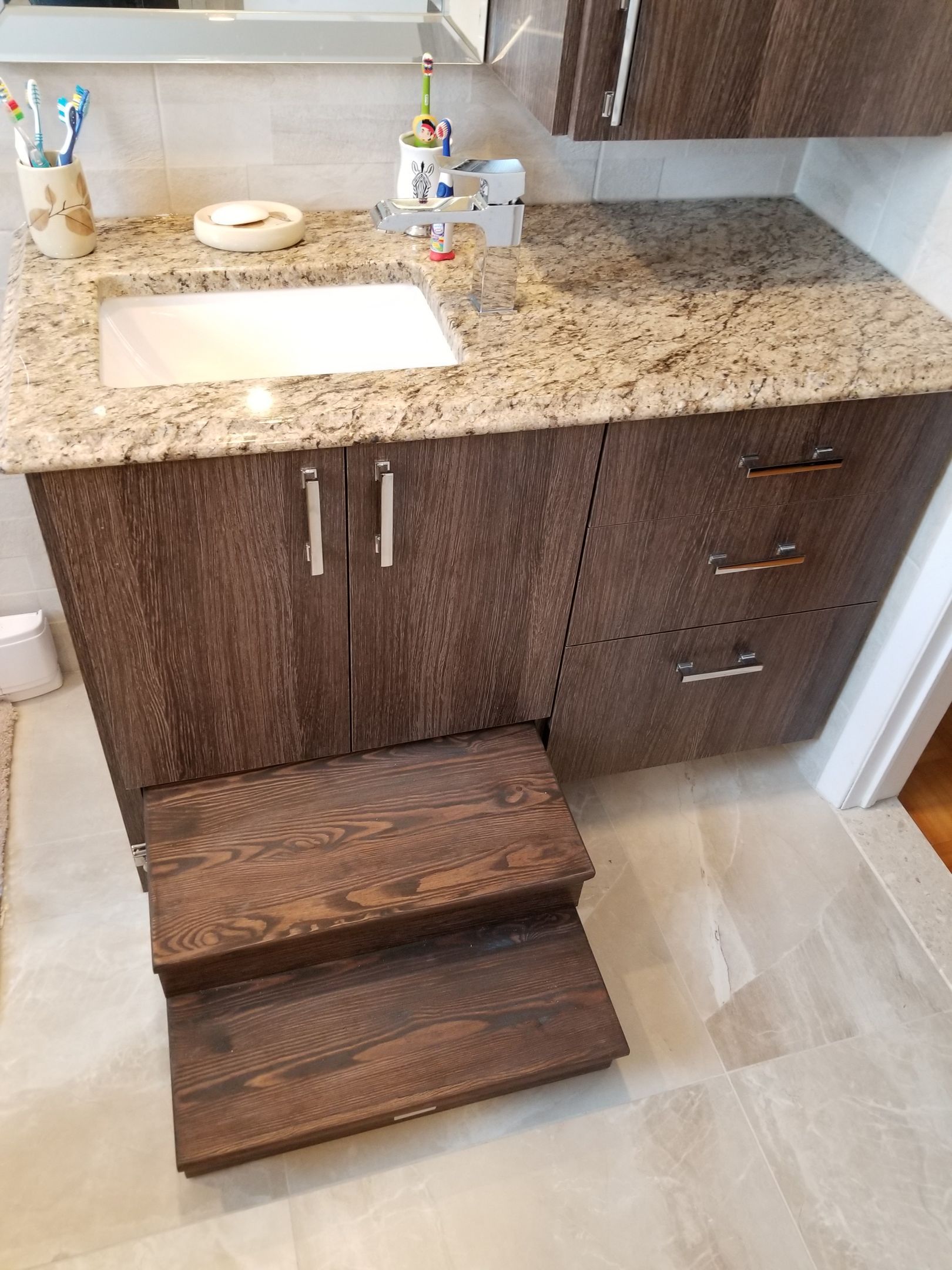 Meuble-lavabo de salle de bain avec armoires en bois foncé, comptoir en granit, lavabo et marchepied intégré.
