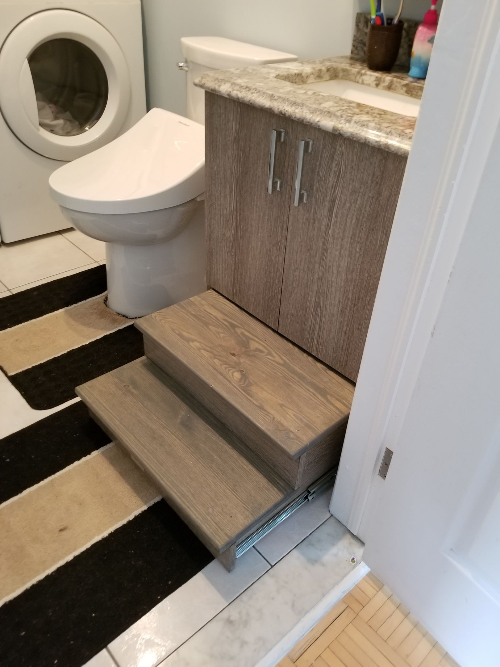 Des marches en bois dans la salle de bains mènent aux toilettes et à un lavabo ; une machine à laver est visible.