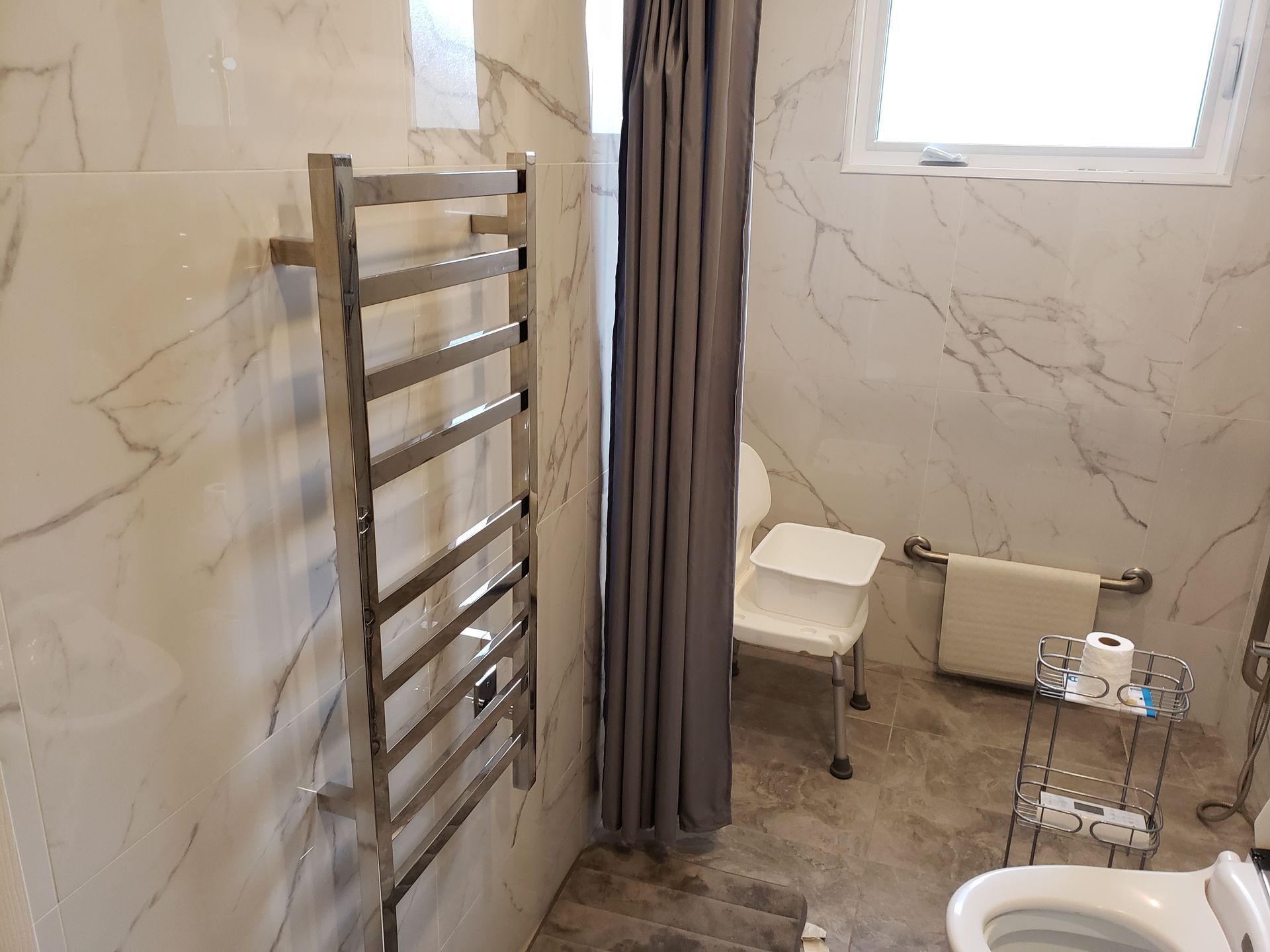 Salle de bains avec sèche-serviettes chauffant, rideau de douche, siège adapté et toilettes. Murs imitation marbre blanc.