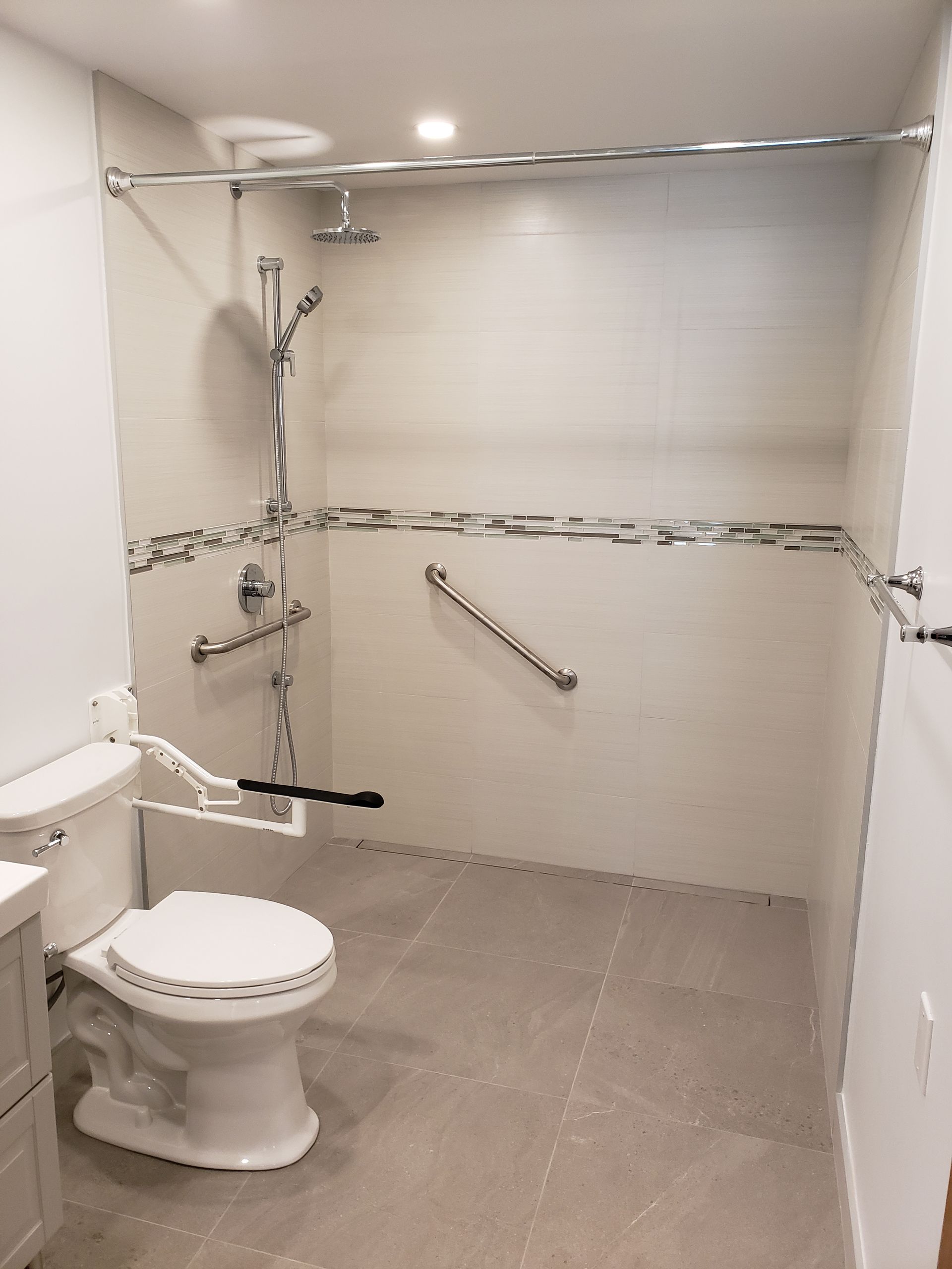 Salle de bain accessible avec toilettes, douche, barres d'appui et carrelage de couleur neutre.