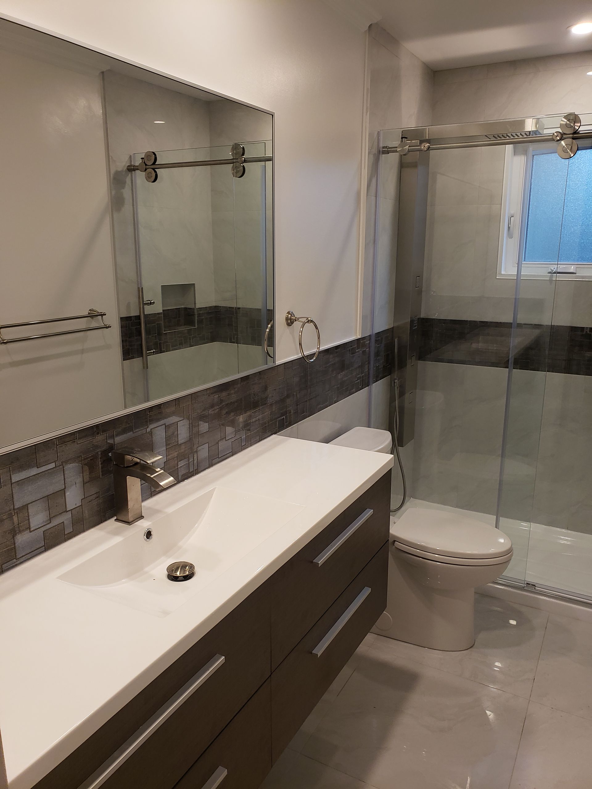 Salle de bains moderne avec comptoir blanc, armoires foncées, douche en verre et mur d'accent en mosaïque.