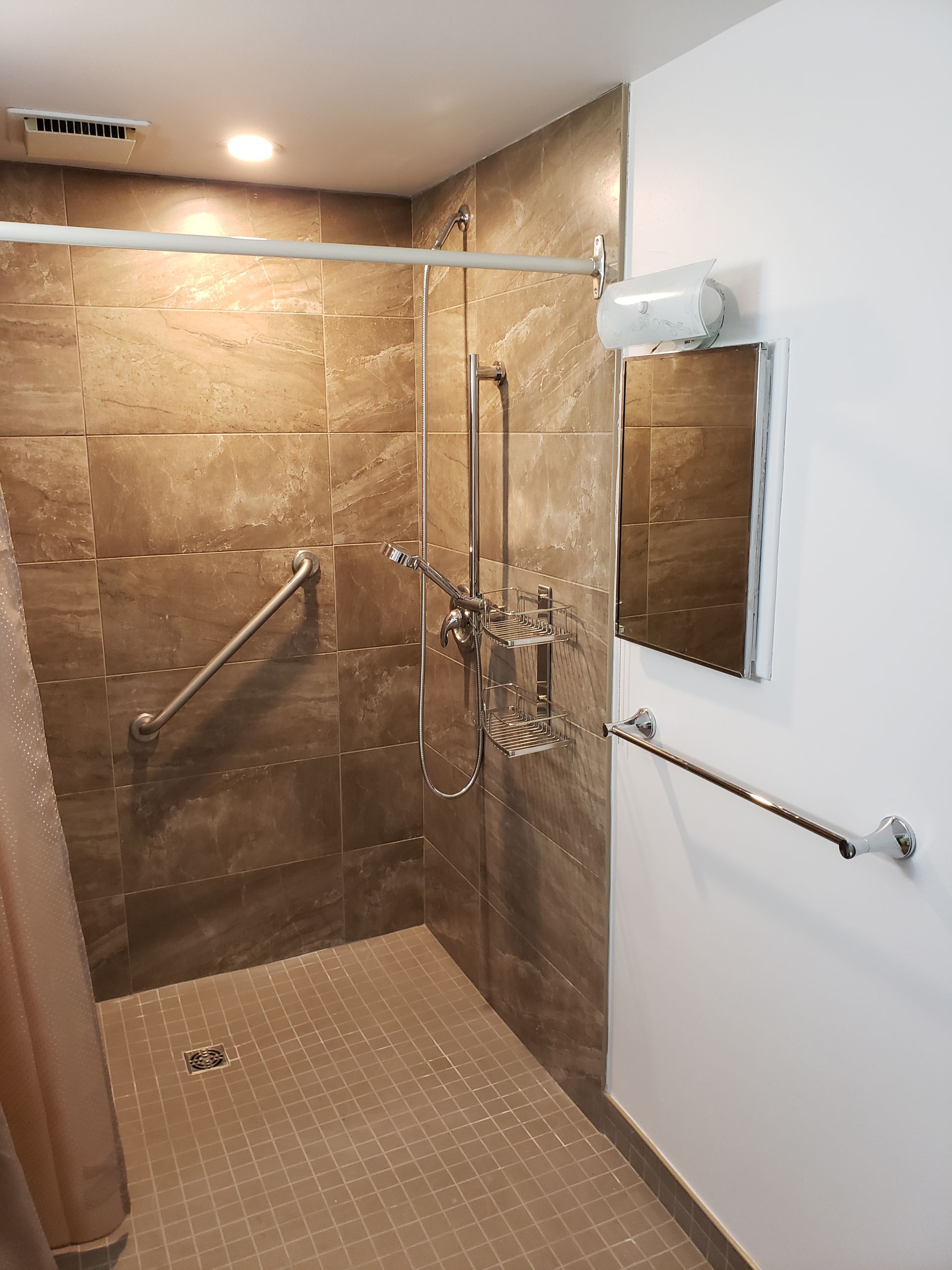 Cabine de douche accessible avec barres d'appui et douchette, carrelage marron et seuil bas.