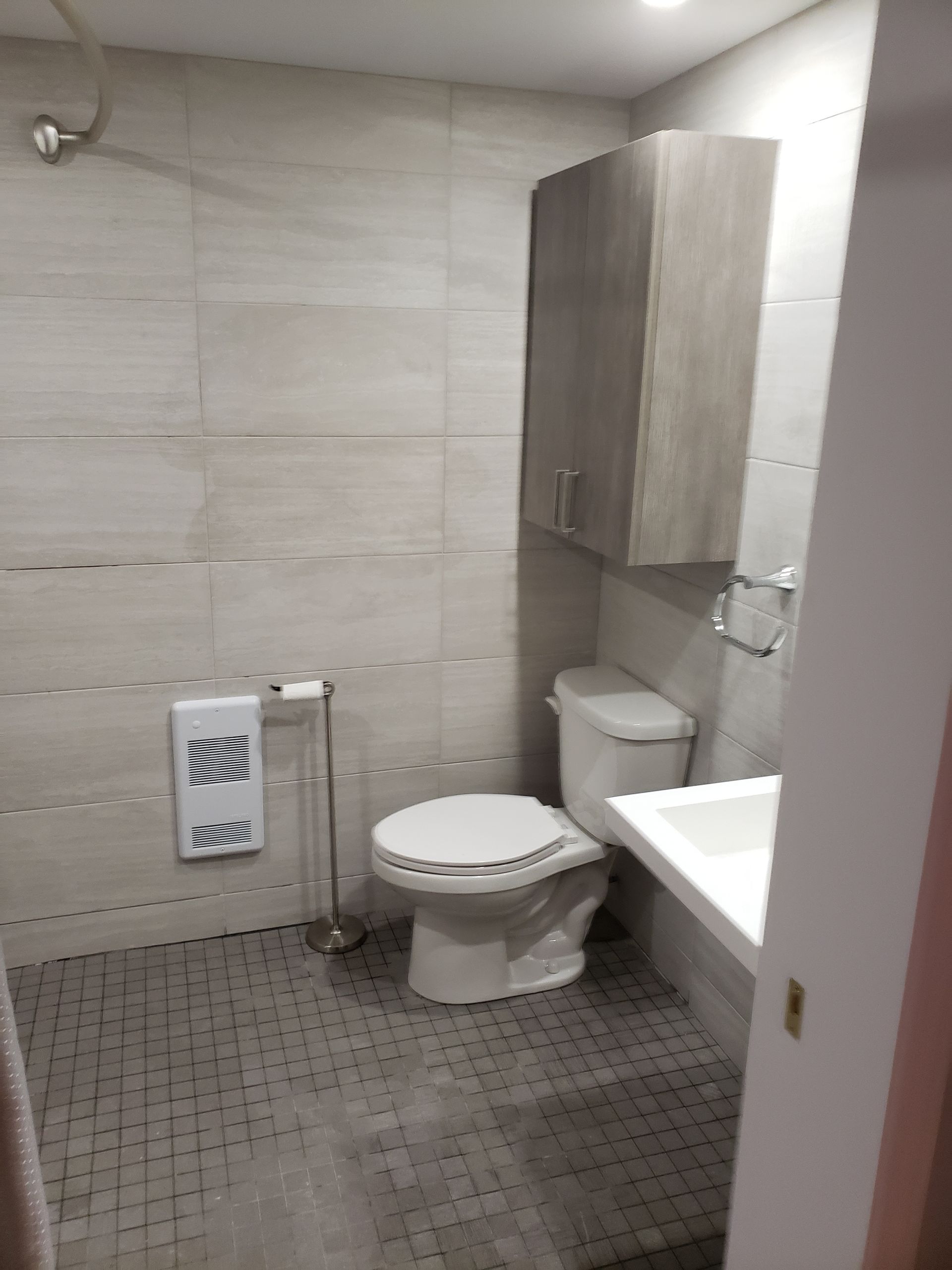 Salle de bains avec murs et sol carrelés gris clair, toilettes, meuble de rangement, lavabo et radiateur mural.